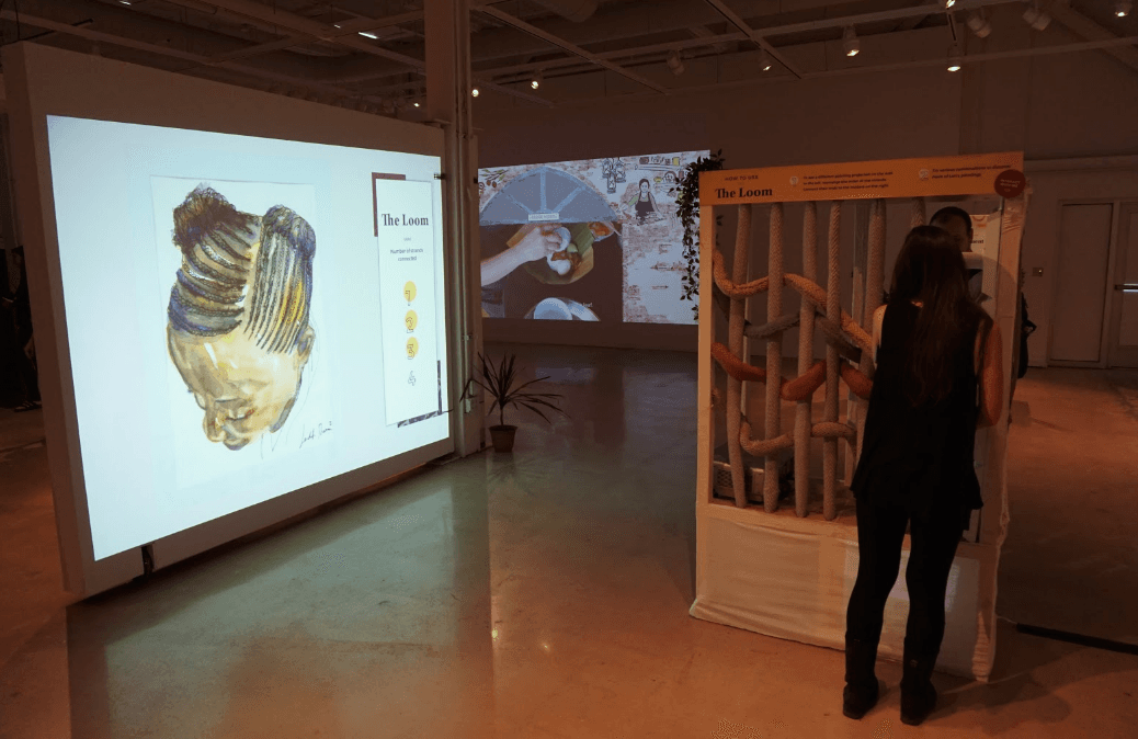 The Loom on display at an exhibit, shown alongside a large projected painting and a visitor rearranging the rope strands